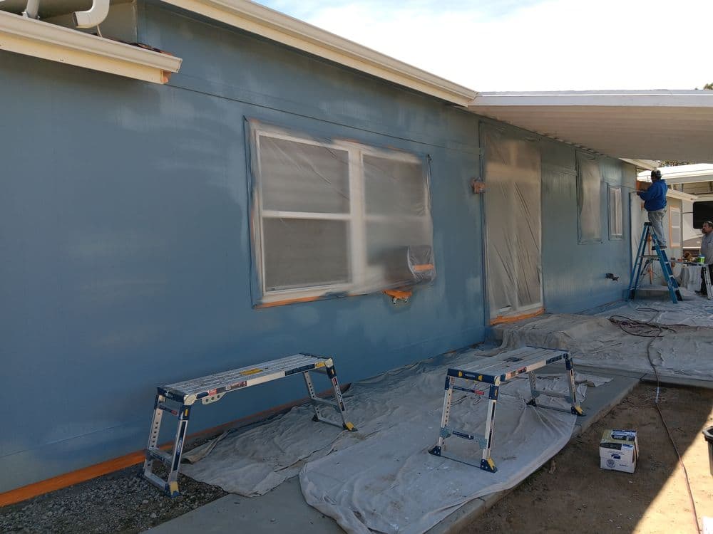Blue house exterior being painted with protective coverings and ladders set up for work.