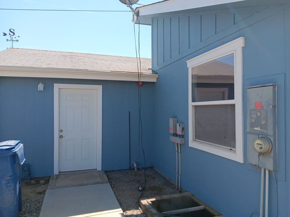 Blue house exterior with side door, utility meter, and nearby trash bins under clear sky.