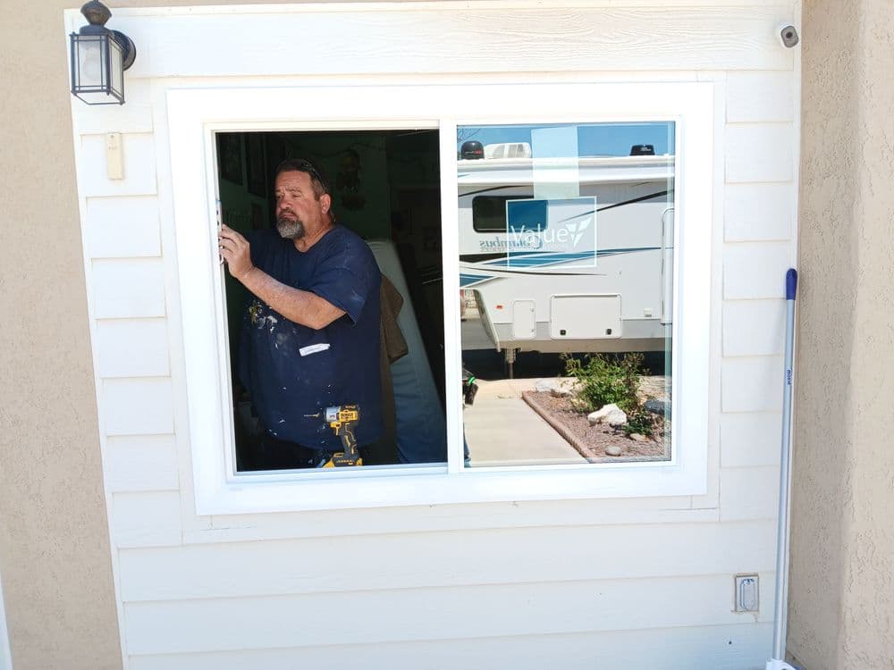 Man installing a window in a home, with a recreational vehicle and garden visible outside.