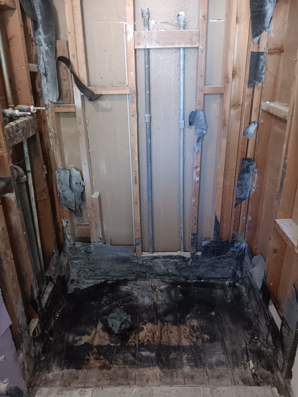 Damaged bathroom walls and floor showing exposed pipes and construction materials.
