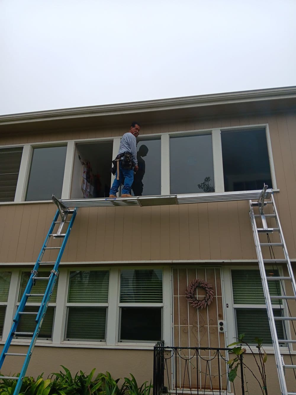 Worker using a ladder to install windows on a beige house exterior. Safety precautions observed.