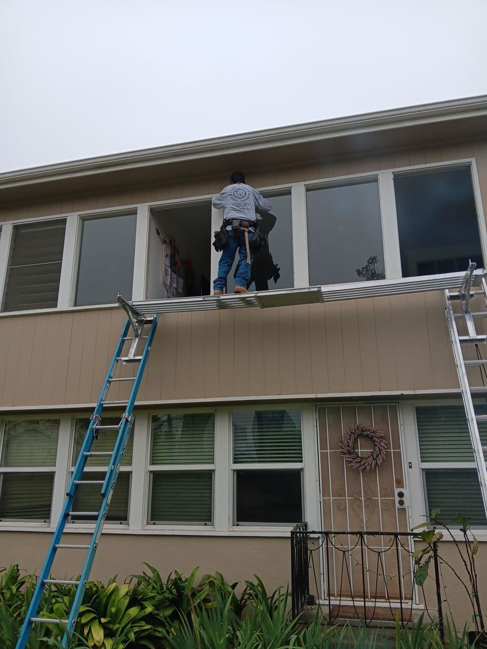 Worker repairing windows on a multi-story house using a ladder for access.