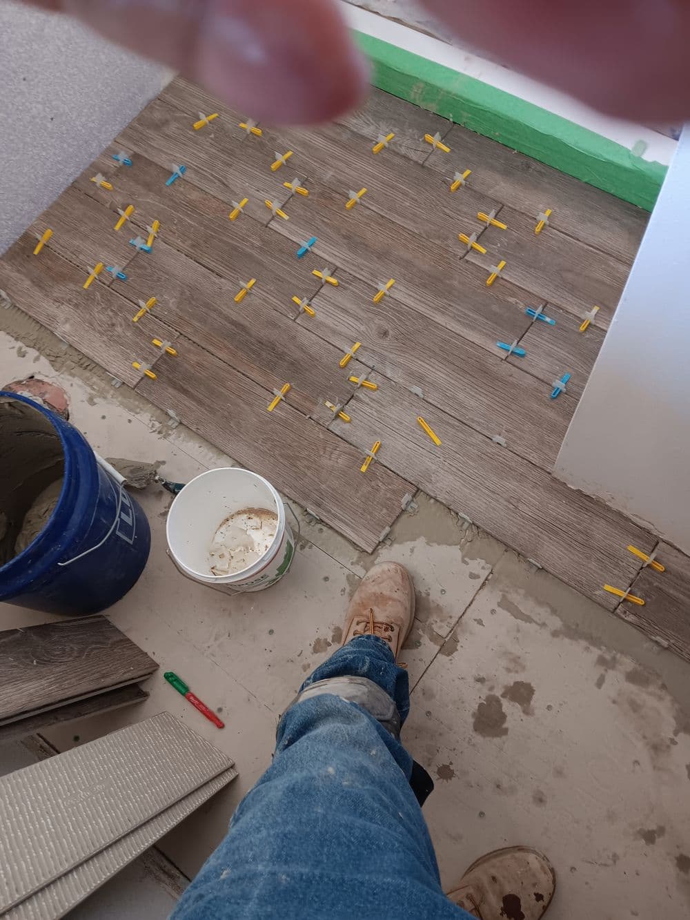 Floor tiling process with spacers, a bucket of grout, and a worker's foot in view.