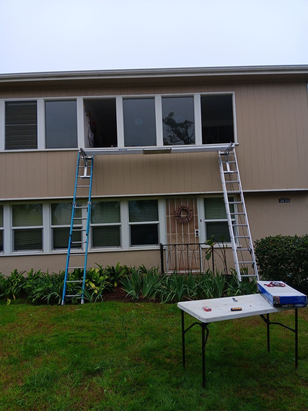 Ladders positioned at a house for window repairs, with tools on a table below.