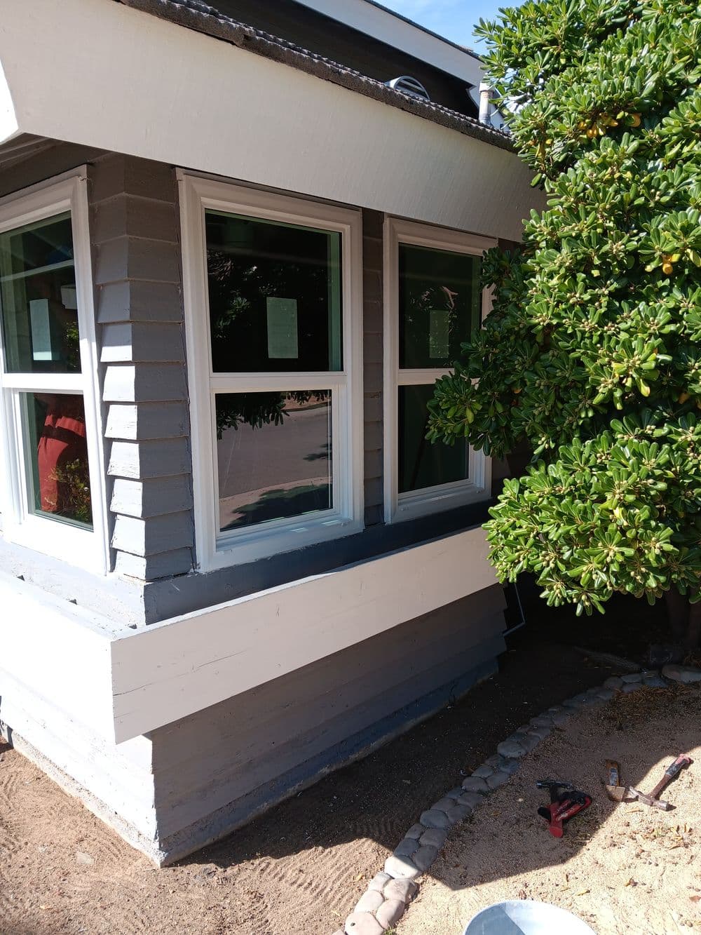Corner of a house with new windows, gray siding, and surrounding greenery. Tools on ground.
