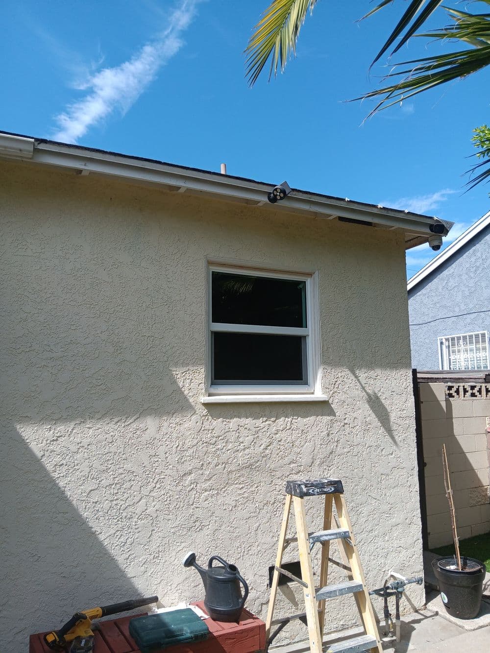Exterior house wall with a window, a ladder, and gardening tools under a clear blue sky.