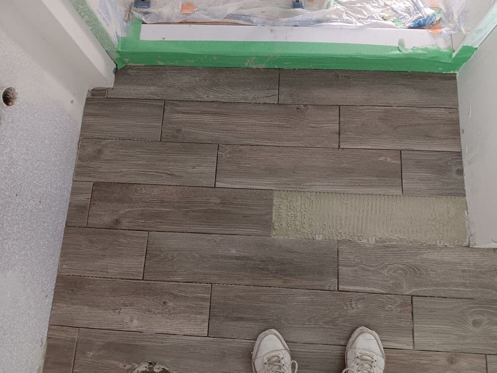 Gray wood-look tile flooring installation in progress, with green masking tape along the wall.